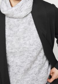 Grauer gestrickter Pullover mit einem Kragen, unter einem schwarzen Blazer getragen. Der Pullover hat ein strukturiertes Muster und wirkt behaglich.