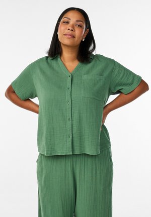 Naine lahtise rohelise nööpidega särgi ja sobivate pükstega seisab valgel taustal käed puusas.