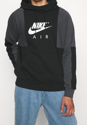 Osoba ubrana w czarną i ciemnoszarą bluzę z kapturem Nike Air z przednią kieszenią i jasnoniebieskie dżinsy, stojąca na białym tle.