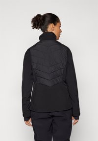 Svart jakke med en tettsittende design og en puffy, quilted bakdel i chevronmønster og høy krage. Lange ermer med tommelhull.