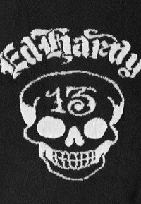 Zwarte stof met een witte gebreide ontwerp van een schedel en de tekst "Lil' Hardy" erboven, met opvallende kenmerken zoals holle ogen en een grijnzende mond.