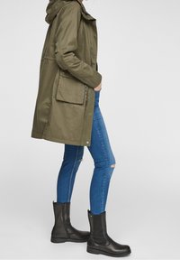 Giacca lunga verde oliva con cappuccio, tasche laterali, abbinata a jeans skinny blu e stivaletti neri.