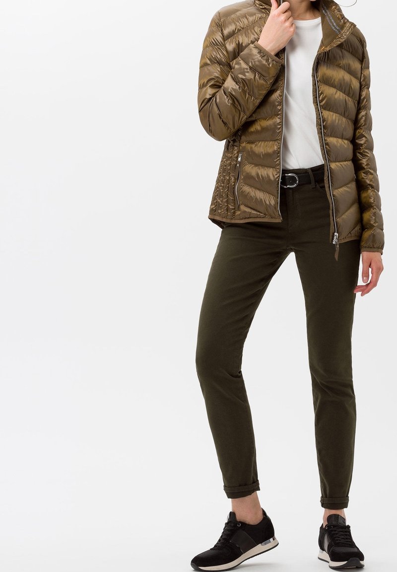 Bronsfärgad puffjacka med quiltad textur, dragkedja fram, ihop med mörkgröna byxor och svarta sneakers. Enkel vit t-shirt under.