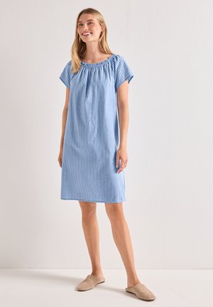 Femme souriante, debout, portant une robe ample à rayures verticales bleues et blanches avec des manches courtes et des chaussons beiges, fond clair uni.