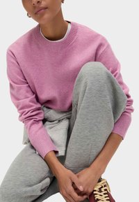 Maglione rosa chiaro lavorato a maglia con colletto a girocollo e polsini a costine, abbinato a pantaloni della tuta grigi. Il modello è seduto, con le braccia appoggiate sulle ginocchia.