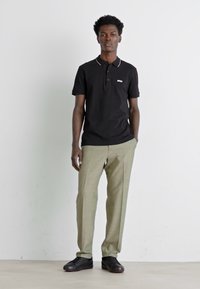 Svart polo skjorte med krage, kortermet, og hvit logo. Lys grønn bukse med skreddersydd passform. Svart sneakers. Nøytral bakgrunn.