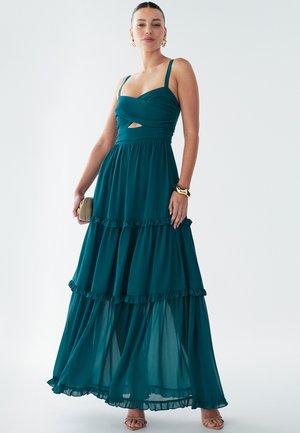 Tyrkysové maxi šaty s volánkovým vrstveným lemem, s twistovaným korzetem a výřezem v pase. Látka vypadá lehce a splývavě.