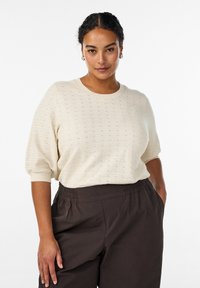 Top de punto texturizado color crema con cuello redondo y mangas cortas y voluminosas. Combinado con pantalones de cintura elástica en marrón oscuro.