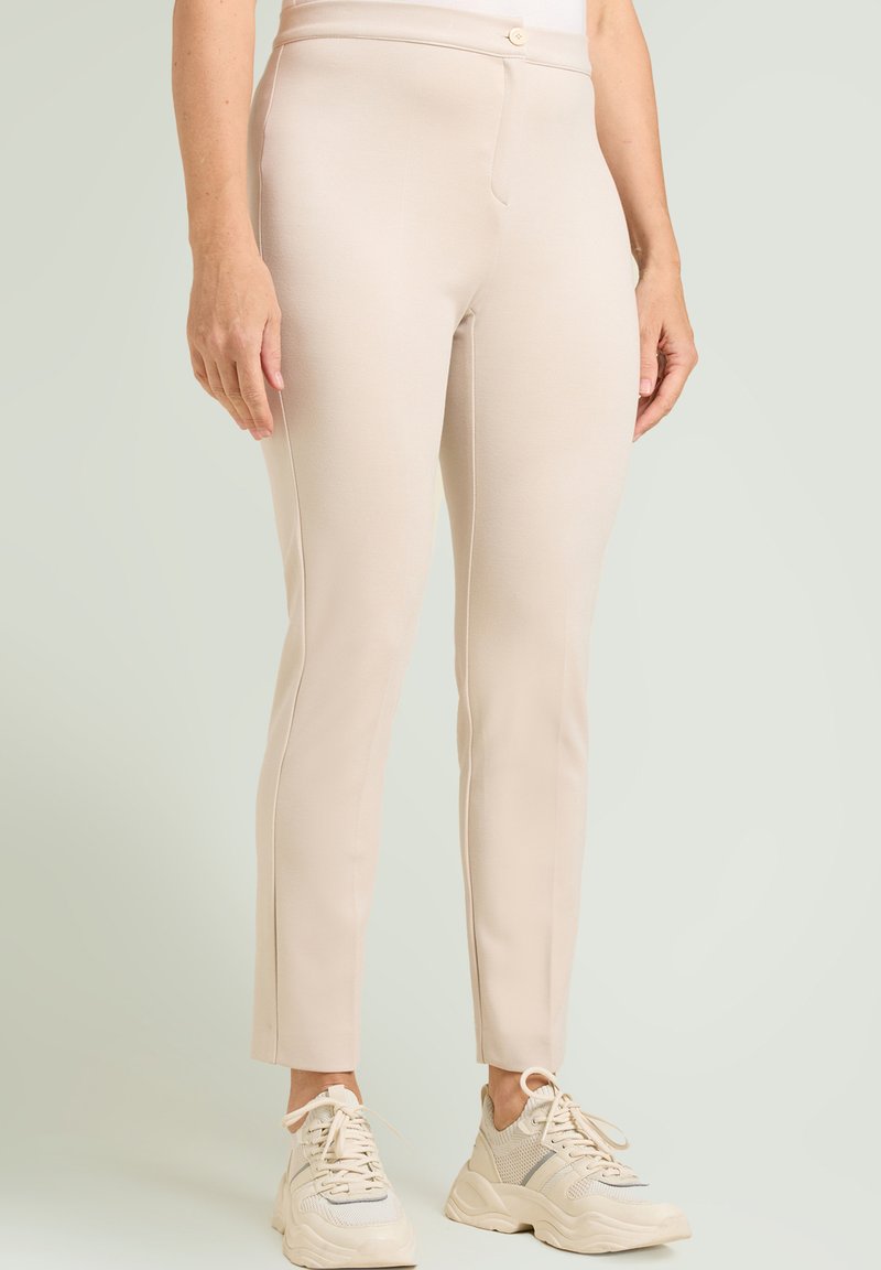Pantalones ajustados beige hechos de tela suave con textura lisa, con cierre frontal de botones y bolsillos laterales, combinados con zapatillas claras.