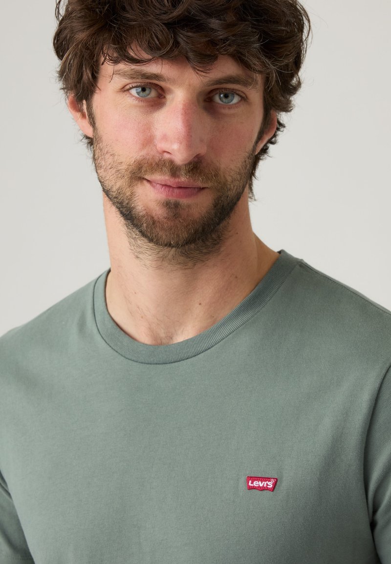 Man with curly brown hair and beard wearing a sage green Levi's t-shirt against a plain light background.