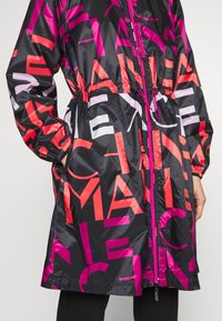 Černá lehká bunda s velkými abstraktními písmeny v jasně růžové, červené a levandulové barvě, elastickými manžetami, předním zipem a staženým pasem.