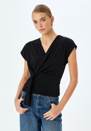 Top nero a maniche corte con design avvolgente e dettaglio del nodo frontale. Tessuto liscio e elastico in una silhouette aderente abbinato a jeans blu.