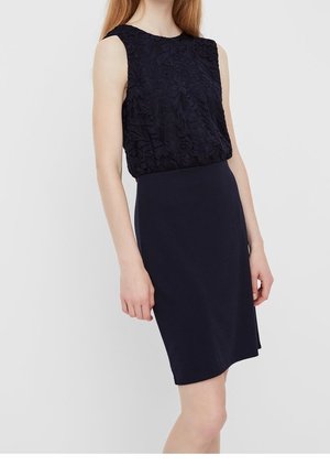 Vrouw die een mouwloze zwarte jurk draagt met een getextureerd bloemenkant bovenstuk en een gladde, nauwsluitende rok, staand tegen een eenvoudige lichte achtergrond.