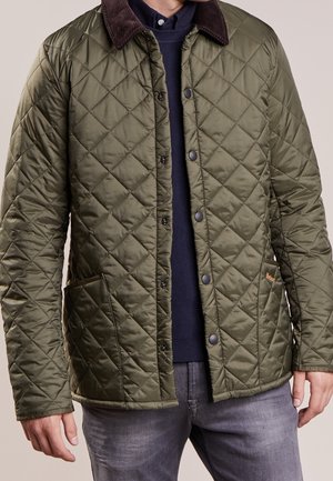 Mann trägt olivgrüne gesteppte Jacke mit dunkelbraunem Kragen, marineblauen Pullover und graue Jeans, steht vor einfarbigem Hintergrund.