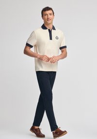 Beige polo tröja med marinblå krage och manschetter, utrustad med en logotyp på vänster bröst. Bärs med mörka byxor och bruna skor.