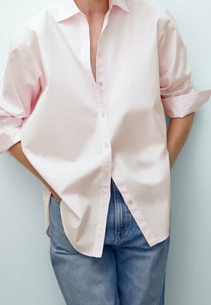 Person wearing a light pink button-up shirt with rolled-up sleeves and blue jeans, standing with hands in pockets against a light background.