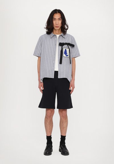 Chemise à manches courtes rayée bleu clair et blanc avec un accent nœud noir, associée à un short foncé. Design notable avec poche avant.