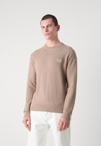 Maglione lavorato a maglia beige con scollatura a girocollo e polsini e orlo a costine. Presenta una piccola etichetta con il marchio sul petto.