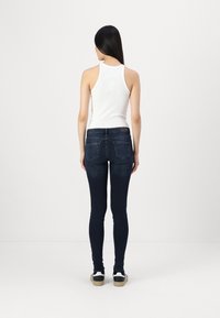 Fehér bordás ujjatlan felső sötétkék skinny farmernadrággal párosítva; jellemzője a normál hátsó zsebek és a minimális varrás; napi viseletként látható lábbeli.
