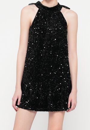 Robe noire sans manches à paillettes, style dos nu, col haut avec des rubans noués aux épaules, portée par une personne debout devant un fond uni.