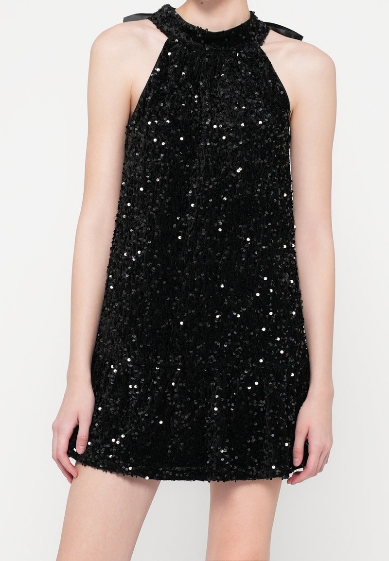 Robe noire sans manches à paillettes, style dos nu, col haut avec des rubans noués aux épaules, portée par une personne debout devant un fond uni.