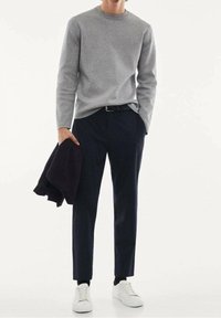 Suéter gris, pantalones oscuros de color marino y zapatillas blancas. El suéter tiene cuello redondo; los pantalones son ajustados. Sostiene una chaqueta oscura en una mano.