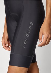 Sorte sykkelshorts laget av elastisk materiale, med en strømlinjeformet design, flatlås sømmer, og "Isadore" trykket vertikalt i grått.