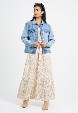 Een lichtblauwe denimjack met een klassieke pasvorm, voorzien van twee voorzakken en een knoopsluiting, gedragen over een gele bloemenmaxi-jurk en beige laarzen.