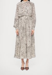 Femme portant une robe blanche à manches longues et col haut avec un motif de points noirs irréguliers et des talons ouverts marron.