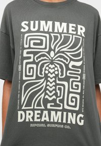T-shirt gris avec impression graphique blanche "SUMMER DREAMING" de motifs abstraits et de designs de feuilles. Matière en coton, coupe décontractée.