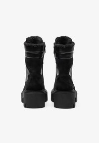 Schwarze Knöchelstiefel mit einem plüschigen, mit Fell gefütterten Schaft, bestehend aus einer Mischung aus Wildleder und Leder, sowie dicken Gummisohlen mit vertikalen Rillen.