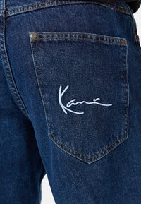 Bolsa trasera de unos vaqueros de mezclilla azul oscuro con costuras naranjas y el logotipo bordado en blanco "Kani" en la bolsa.