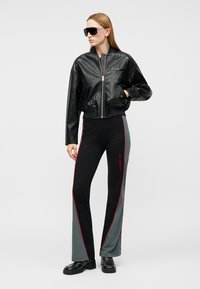 Žena stojaca, oblečená v nadrozmernej čiernej koženej bomber bunde s textúrou, čiernych a sivých nohaviciach s červeným lemovaním, čiernych slnečných okuliaroch a čiernych topánkach.