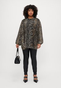 Haut oversized en imprimé léopard noir et beige, associé à un pantalon noir moulant. Les accessoires comprennent un petit sac à main noir et des chaussures à talons avec lanières.