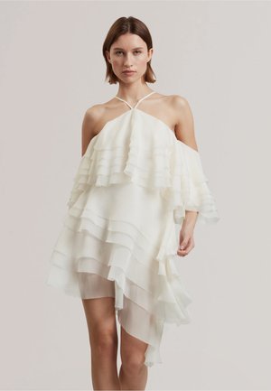 Donna che indossa un abito bianco a strati con spalle scoperte, volant e sottili spalline a halter, in piedi davanti a uno sfondo chiaro uniforme.