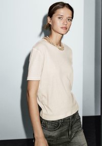 T-shirt beige a maniche corte con scollatura tonda, abbinato a jeans grigi scuri. Tessuto morbido e dettagli di cucitura sottili.