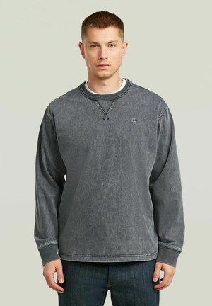 Giovane uomo con capelli corti che indossa una felpa a maniche lunghe nera sbiadita e jeans scuri, in piedi contro uno sfondo chiaro e uniforme.