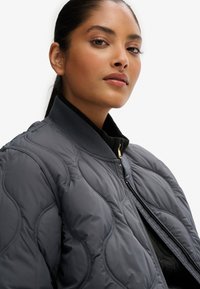 Giacca bomber trapuntata grigia scura con colletto a costine e zip dorata. Presenta una texture liscia con dettagli di cucitura curvi.