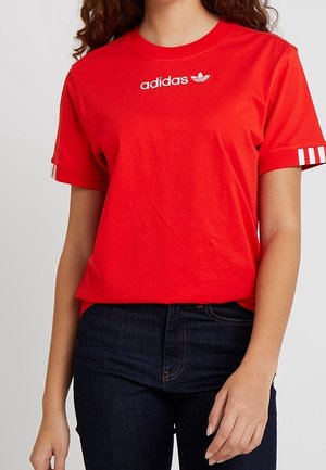 Kobieta w jasnoczerwonym T-shircie Adidas z białym logo i paskami na rękawach, w połączeniu z ciemnoniebieskimi dżinsami, na jednolitym tle.
