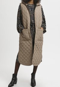 Mujer con un chaleco acolchado largo beige con grandes bolsillos sobre un vestido negro con flores, medias negras y botines negros.