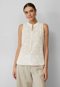 Witte mouwloze blouse met gestructureerde blaadjespatronen, ronde halslijn en een keyhole aan de voorkant. Gecombineerd met lichtbeige broeken.
