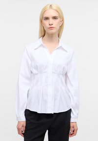 Camicia bianca a maniche lunghe con colletto a punta, caratterizzata da pieghe verticali sul davanti e un orlo curvo, realizzata in tessuto morbido.