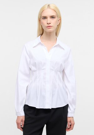 Camicia bianca a maniche lunghe con colletto a punta, caratterizzata da pieghe verticali sul davanti e un orlo curvo, realizzata in tessuto morbido.