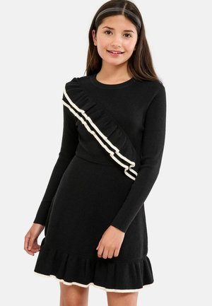 Schwarzes Strickkleid mit langen Ärmeln, das ein diagonales weißes Rüschendetail und einen ausgestellt gesäumten Rüschensaum am unteren Rand aufweist.