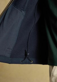 Tumedate jakkide sisemine fleece-vooder, millel on kerimisnööride reguleerimine alumises servas ja nähtav lukuäär vasakul küljel.