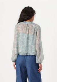 Hellbluse mit floralem Muster und halbtransparenter Textur, mit gerafftem Ausschnitt, langen Ärmeln und tailliertem Design, kombiniert mit dunklen Hosen.
