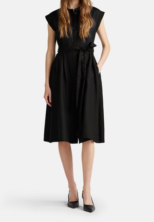 Žena ve černých midi šatech bez rukávů s uvázaným páskem v pase a černých botách s špičatou špičkou, stojící s jednou rukou v kapse.