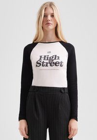 Blonde Frau in einem weißen und schwarzen Langarmshirt mit dem Text "High Street", kombiniert mit hochtaillierten schwarzen Nadelstreifenhosen.