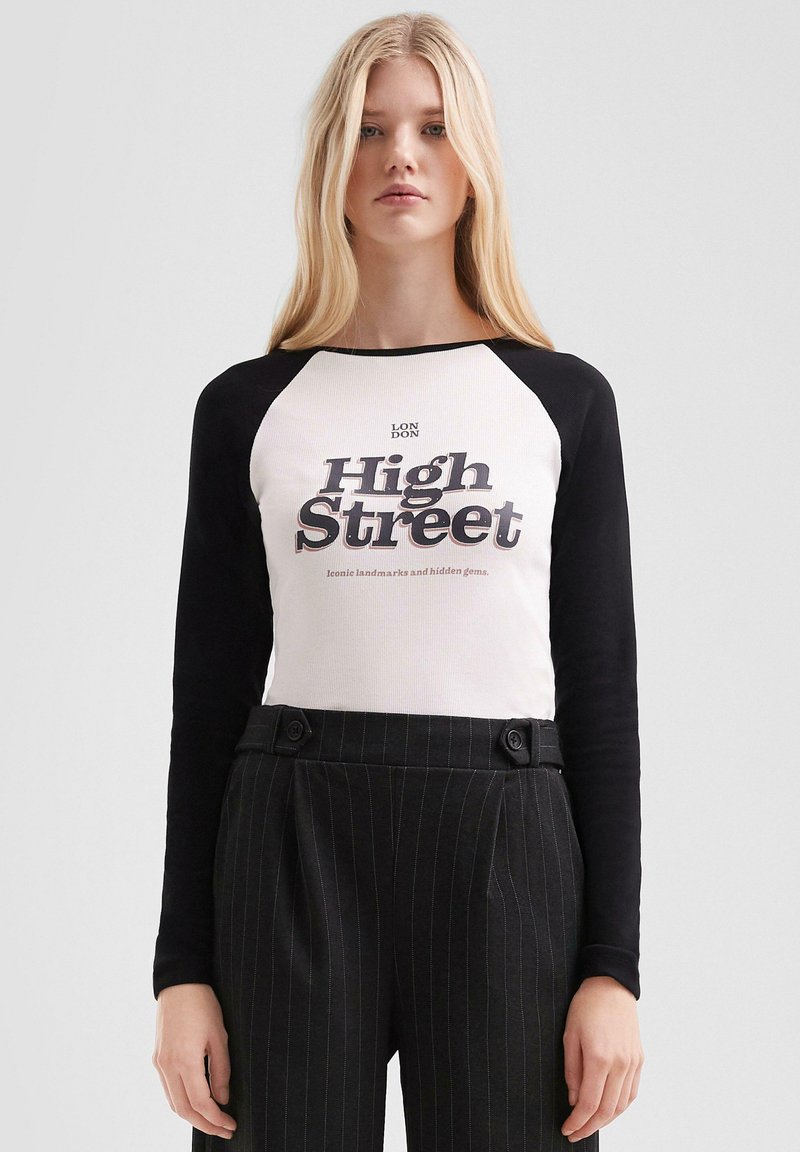 Blonde Frau in einem weißen und schwarzen Langarmshirt mit dem Text "High Street", kombiniert mit hochtaillierten schwarzen Nadelstreifenhosen.