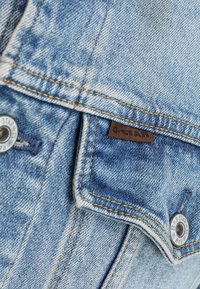 Närbild av en blå denimjackficka med orange sömmar, metal knappar och en liten brun G-Star märke.